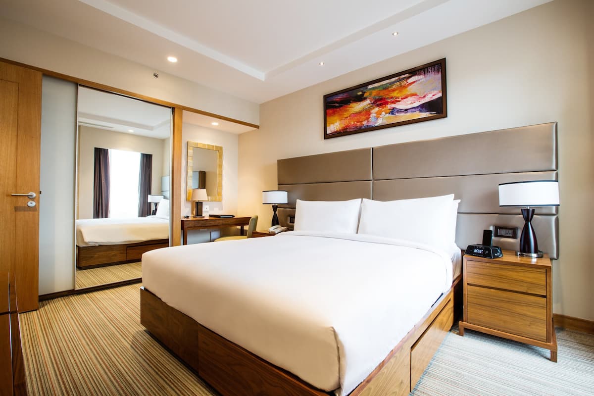 A hotel room with a large bed, a bedside table with a lamp, a nightstand, a mirror, and a painting on the wall.