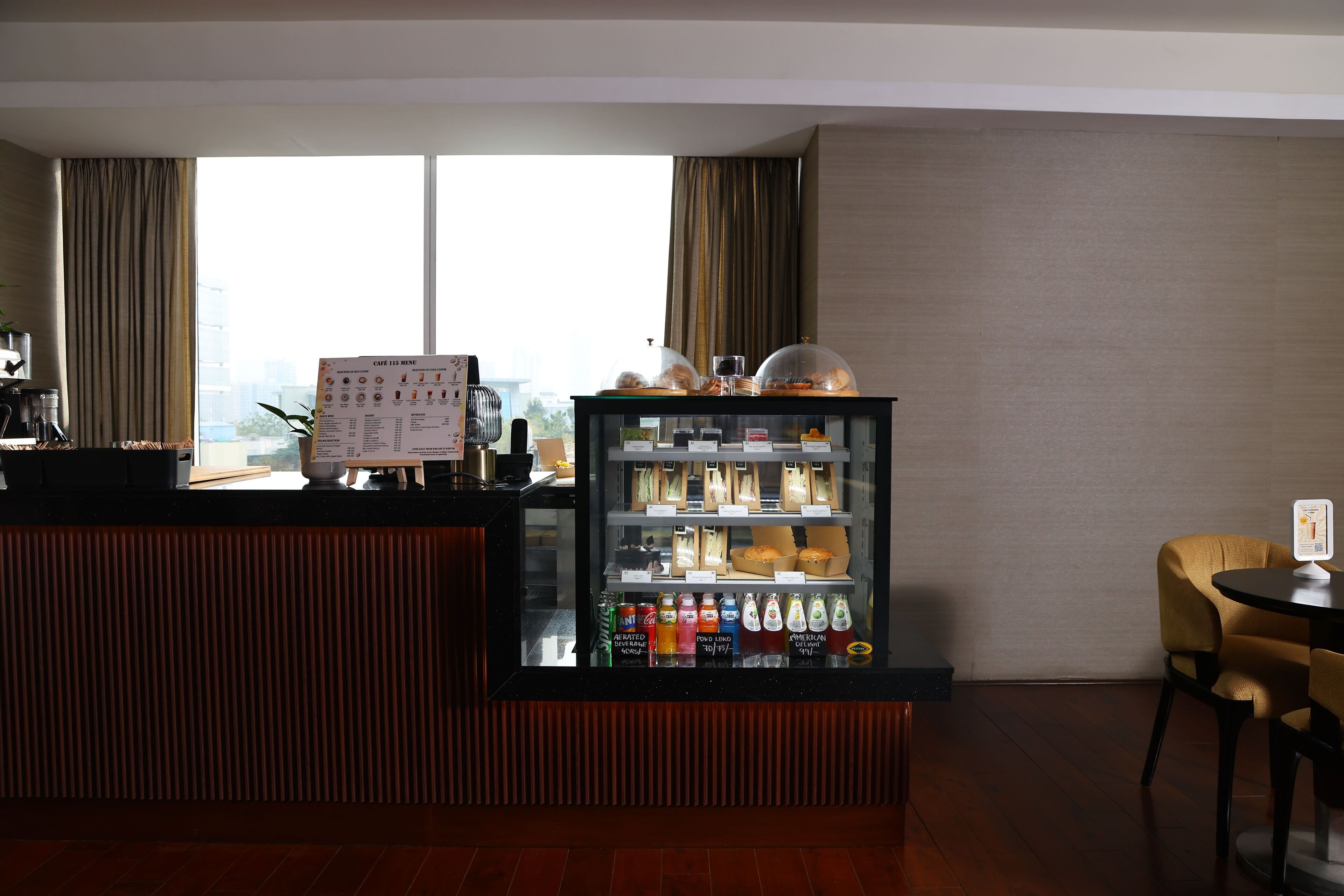 A hotel reception area with a counter, a menu board, a beverage refrigerator, and a view of the city through large windows.