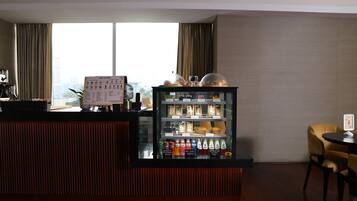 A hotel reception area with a counter, a menu board, a beverage refrigerator, and a view of the city through large windows.