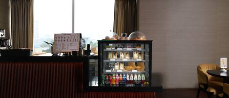 Zona de recepción de hotel con mostrador, cartel de menú, refrigerador para bebidas y vistas a la ciudad a través de amplias ventanas.