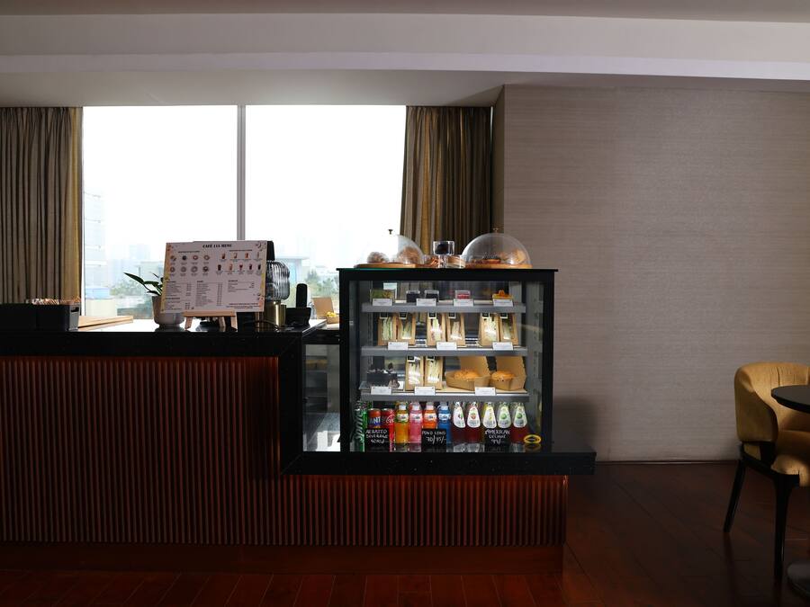 A hotel reception area with a counter, a menu board, a beverage refrigerator, and a view of the city through large windows.