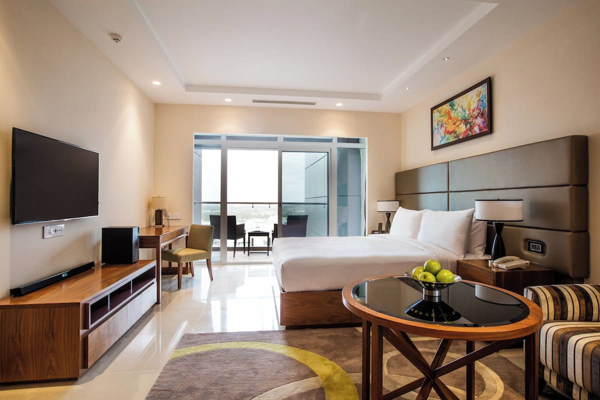 A modern hotel room with a large bed, a flat-screen TV, a dining area, and a balcony with a view.