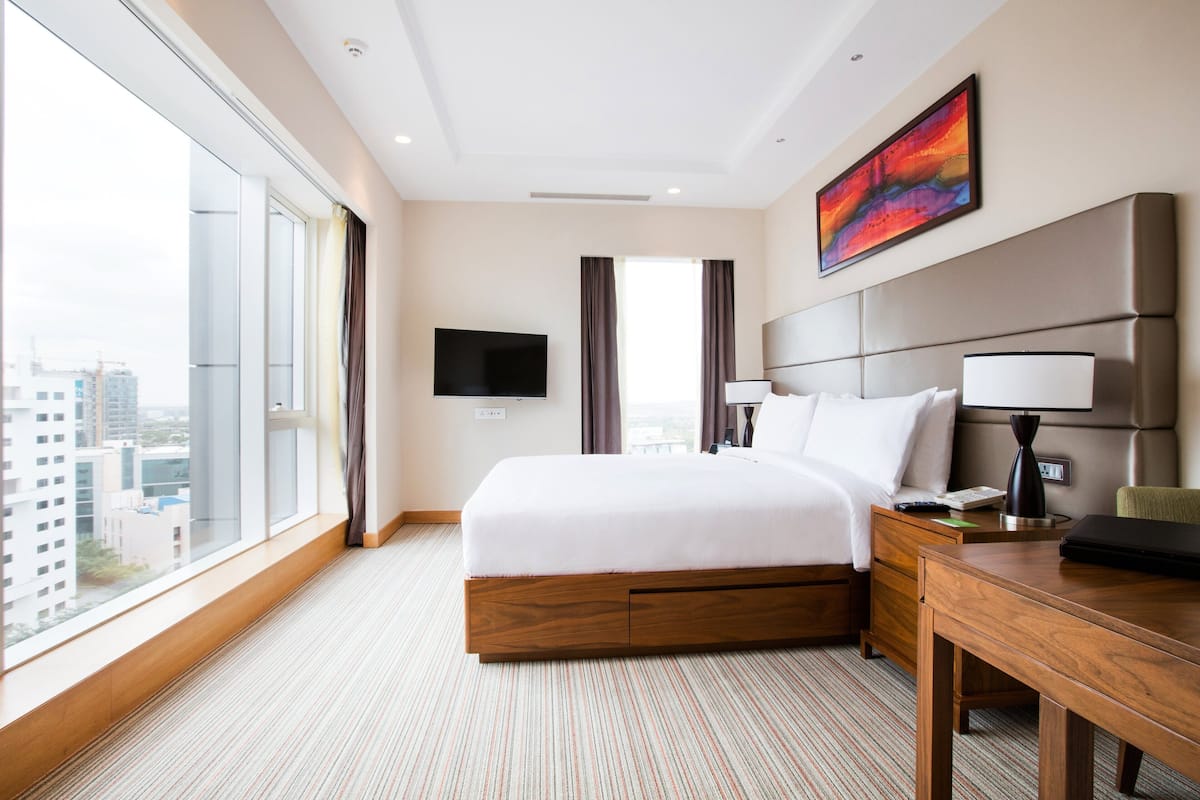 A hotel room with a large bed, a desk, a chair, a flat-screen TV, and a window with a view of buildings.