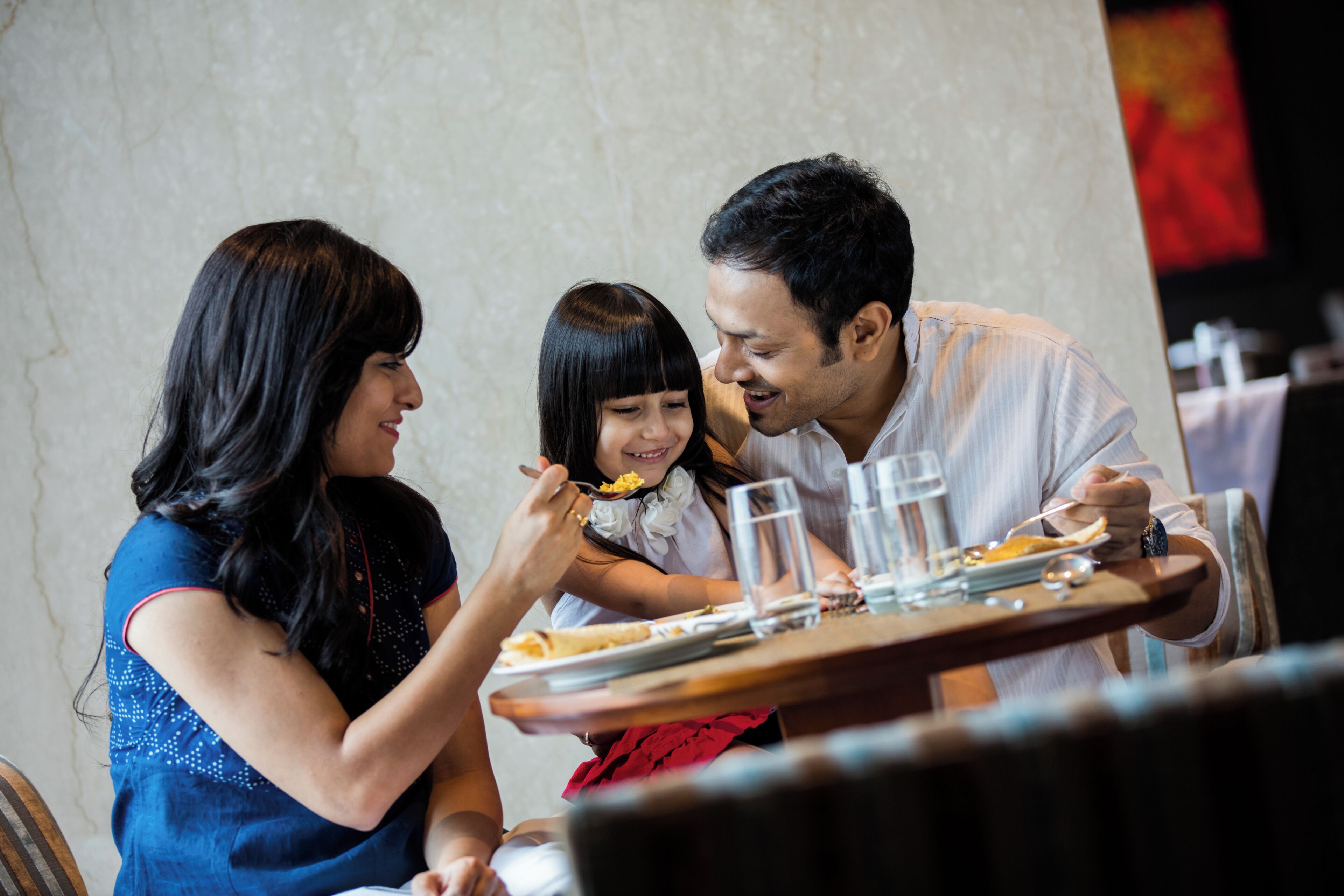 Une famille est assise à une table dans un restaurant. On aperçoit un mur de marbre et, au fond, un drapeau rouge.