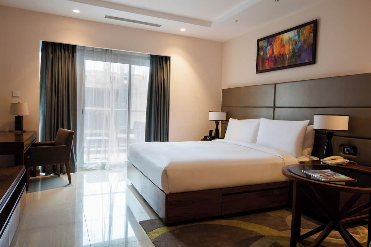 A hotel room with a large bed, a desk, a chair, a bedside table with a lamp, and a painting on the wall.