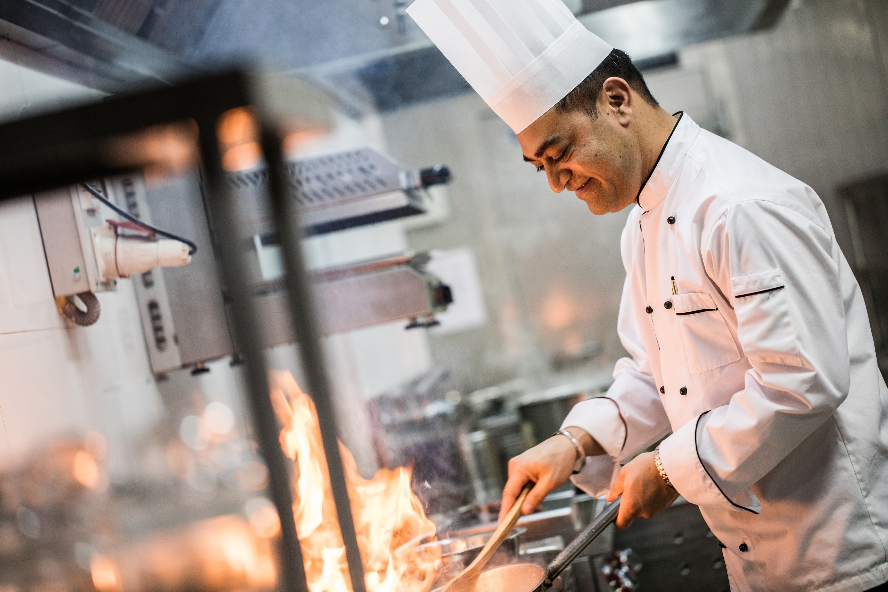 Un chef, vêtu d’un uniforme blanc, cuisine, et on peut voir une grande flamme en arrière-plan.