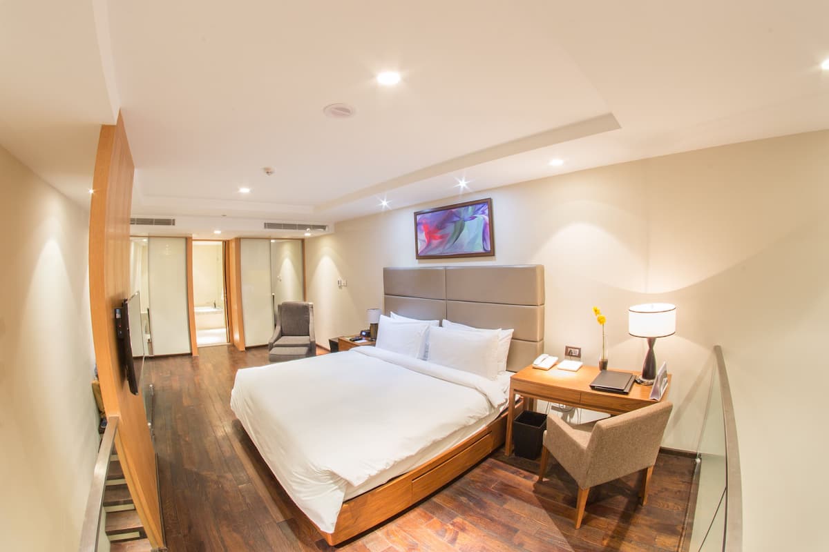 A hotel room with a large bed, a desk with a chair, a small table with a lamp, and a painting on the wall.