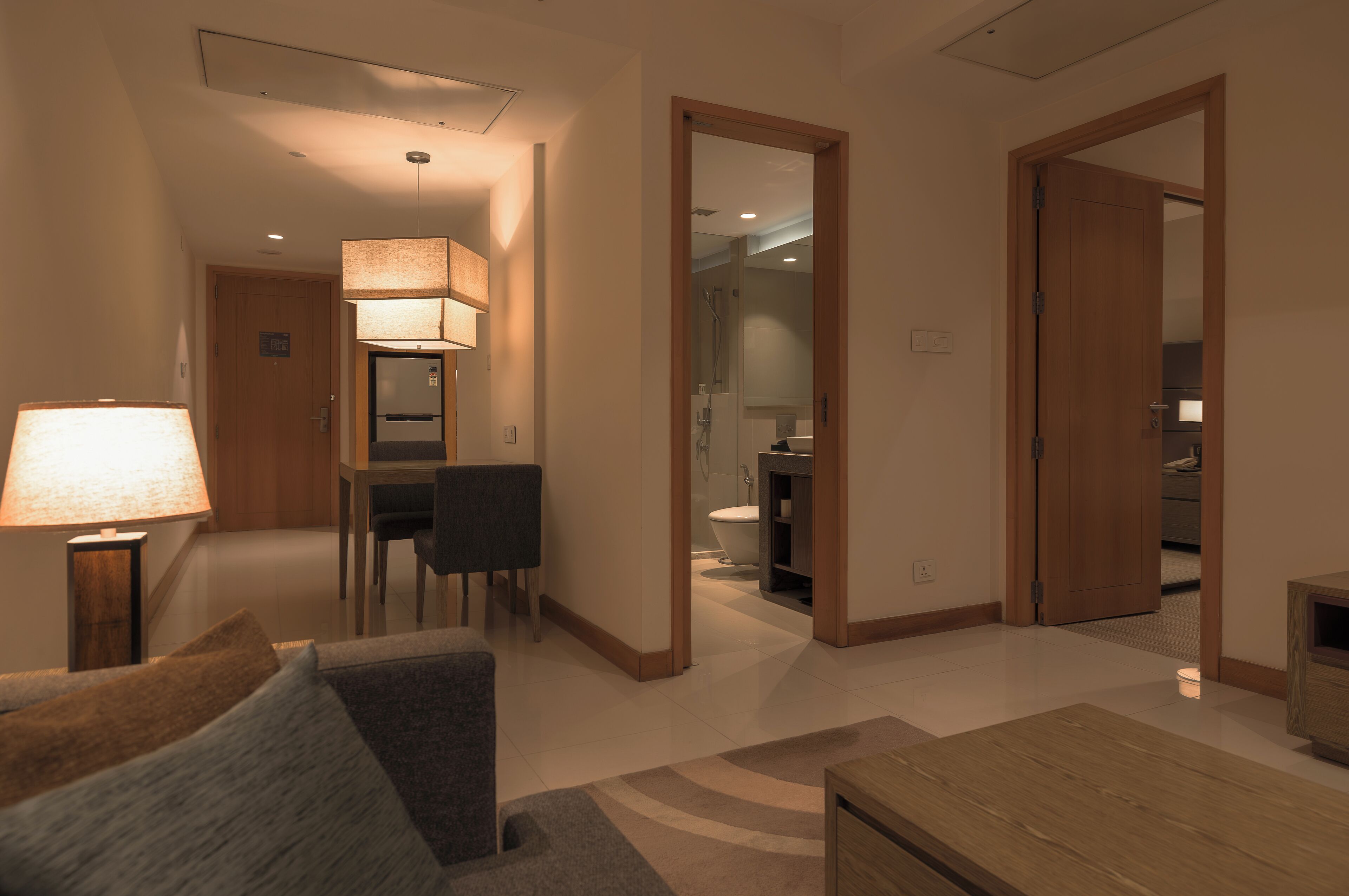 Habitación de hotel con piso de madera, sofá, una mesa y una lámpara. Se ve un baño a través de una puerta abierta.