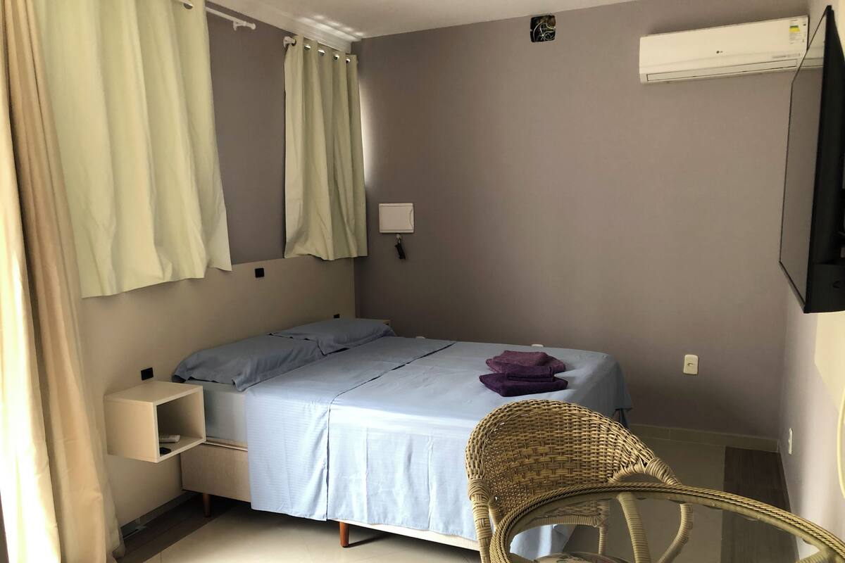 A bedroom with a bed, a nightstand, a wicker chair, and a wall-mounted air conditioner.
