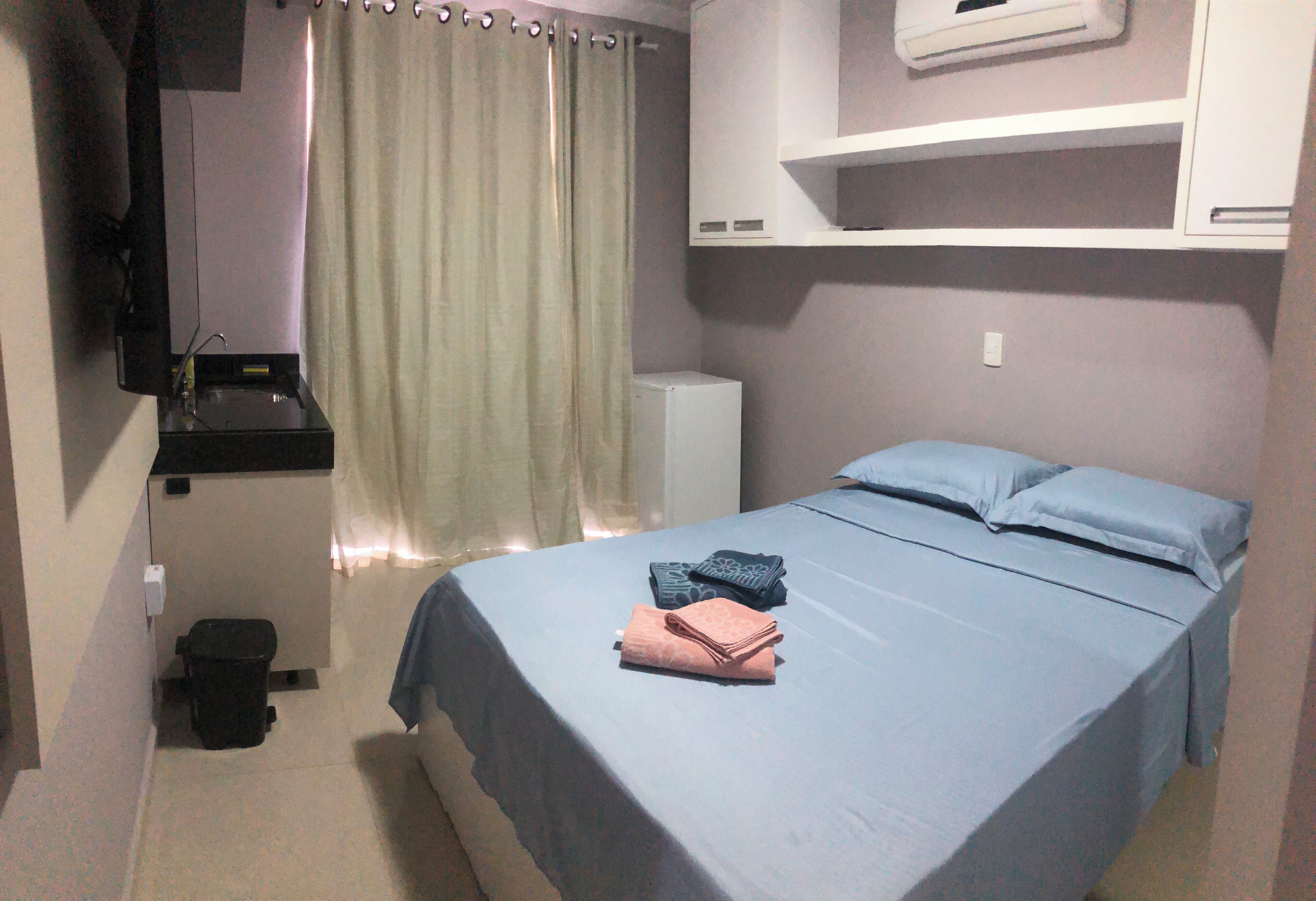 A bedroom with a bed, a small bedside table, a wall-mounted air conditioner, a curtain, and a trash bin.