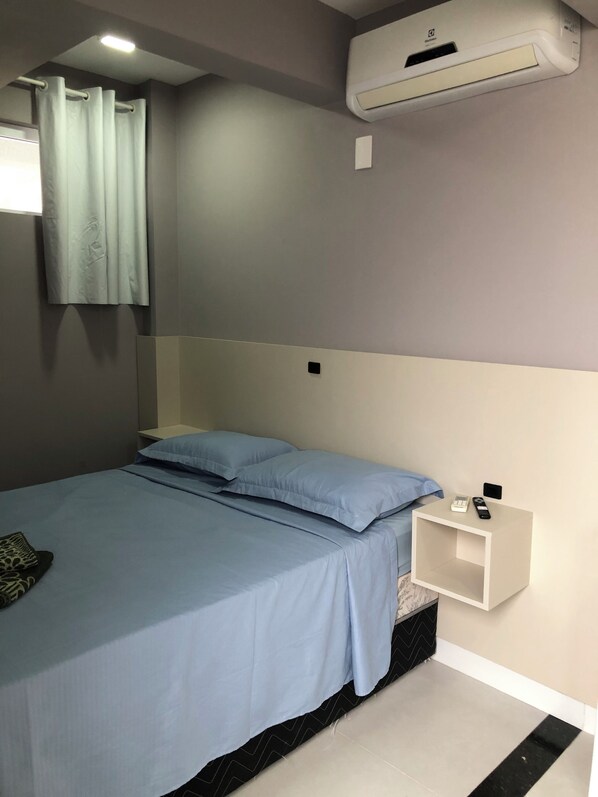 A bedroom with a bed, a nightstand, a wall-mounted air conditioner, and a window with curtains.