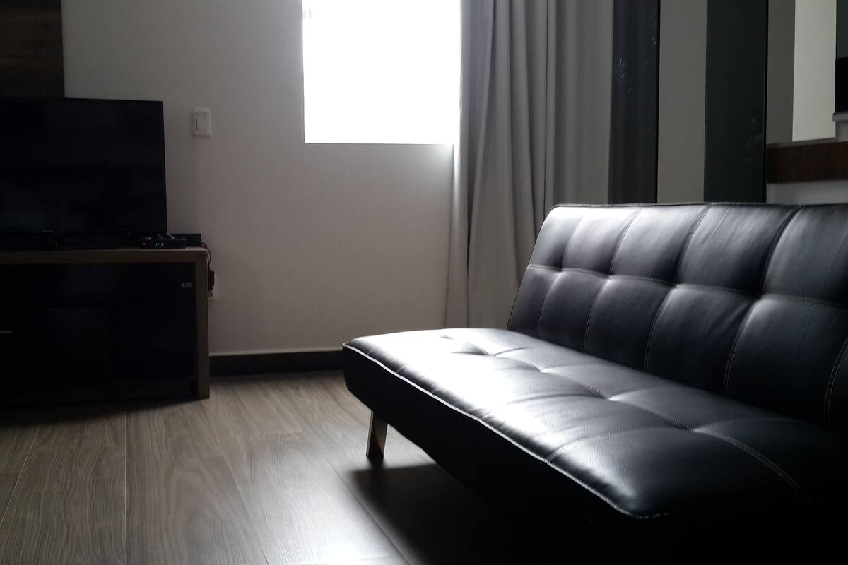 A living room with a black leather sofa, a flat-screen TV on a wooden stand, a window with curtains, and a light-colored wooden floor.