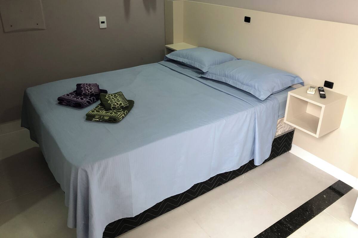 A neatly made bed with a blue bedspread and pillows, a bedside table with a remote control, and a wall-mounted light switch.