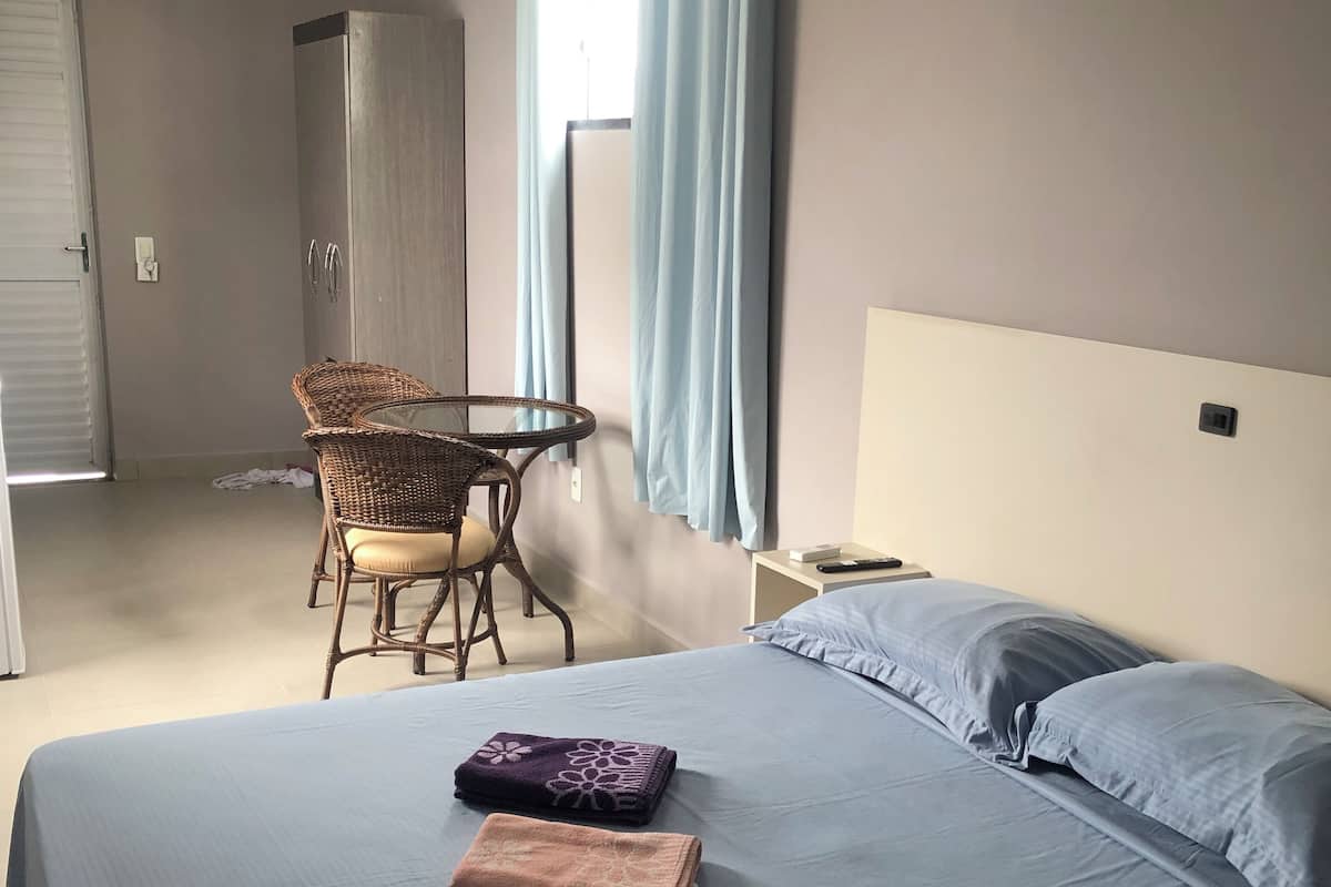 A hotel room with a bed, a chair, a small table, and an air conditioning unit.
