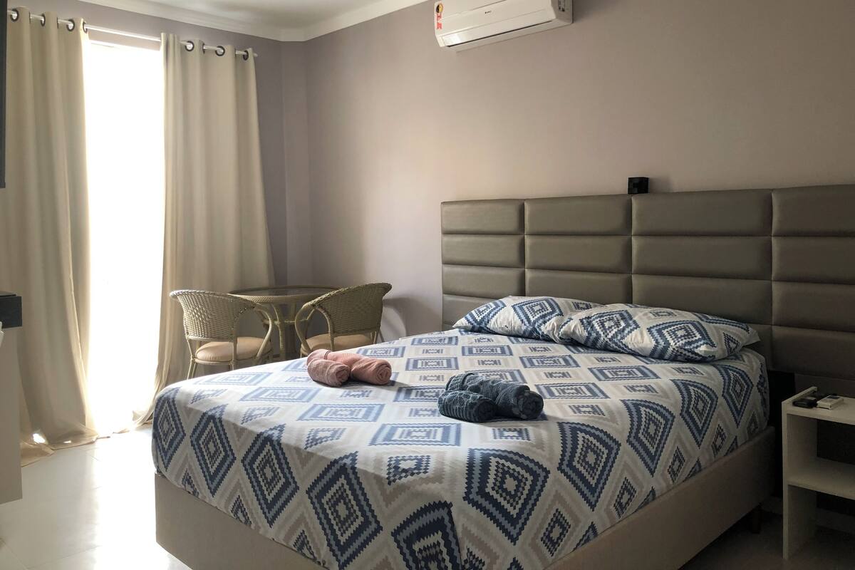 A hotel room with a bed, a small table with chairs, a bedside table with a lamp, and an air conditioning unit on the wall.