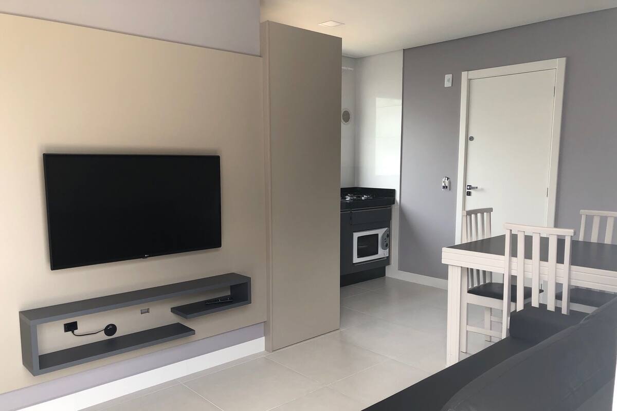 A modern living room with a flat-screen TV, a dining area with a white table and chairs, and a kitchen with a black countertop and oven.
