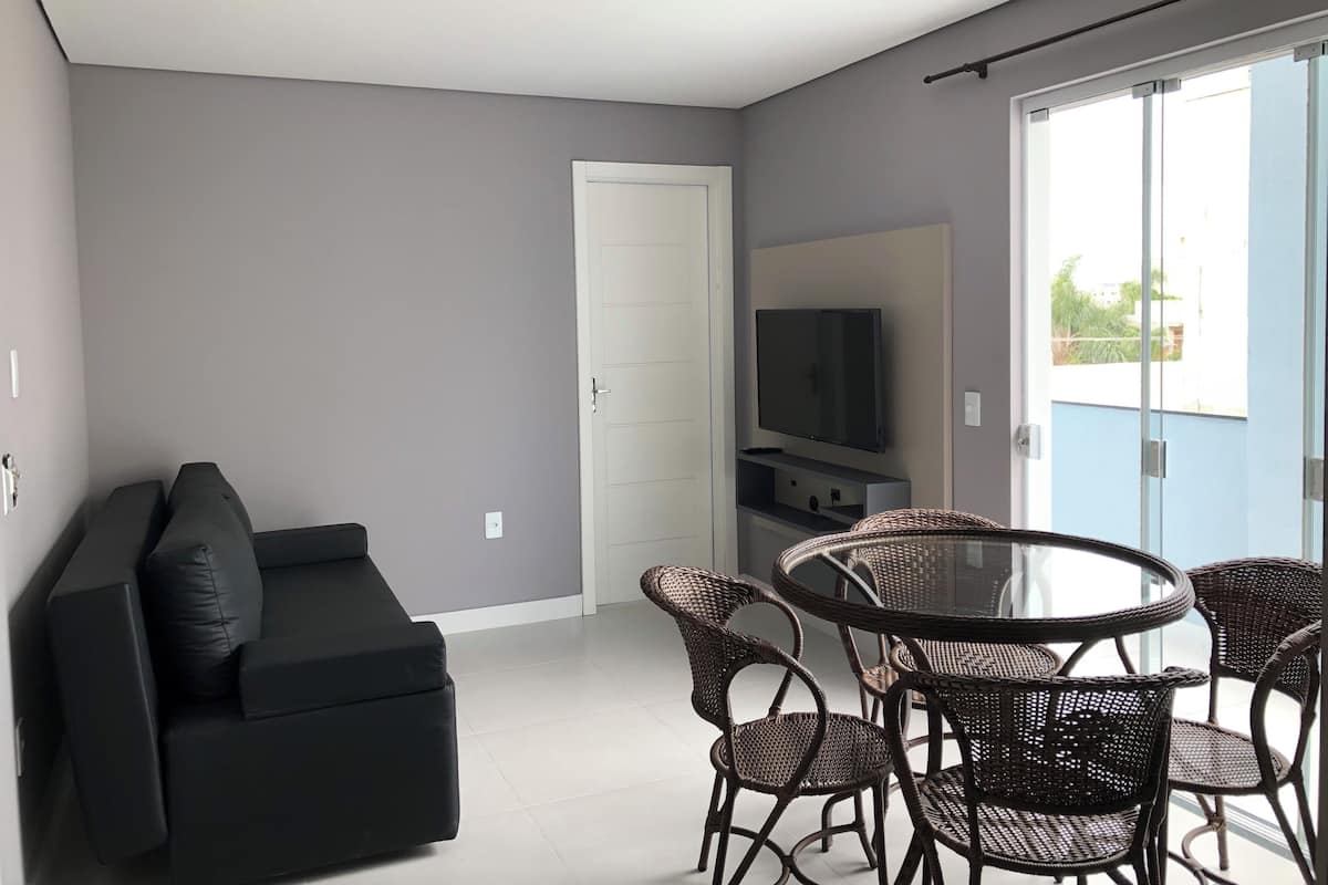 A modern living room with a black sofa, wicker chairs, a flat-screen TV, and a sliding glass door leading to an outdoor balcony.
