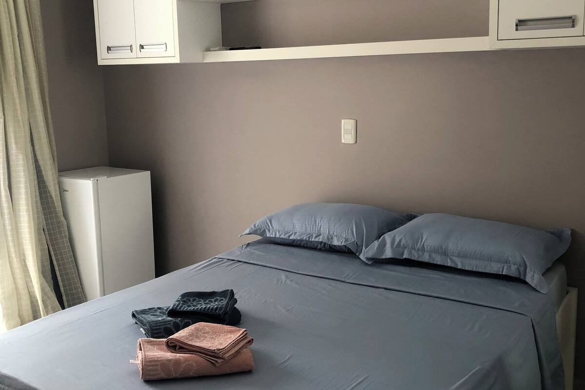 A bedroom with a bed, pillows, a bedside table, a wardrobe, and an air conditioning unit.