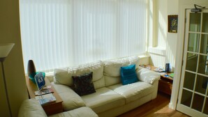 A living room with a beige sofa, two blue pillows, a small table with a lamp, and a window with blinds. - Arlingtons (Blackpool)