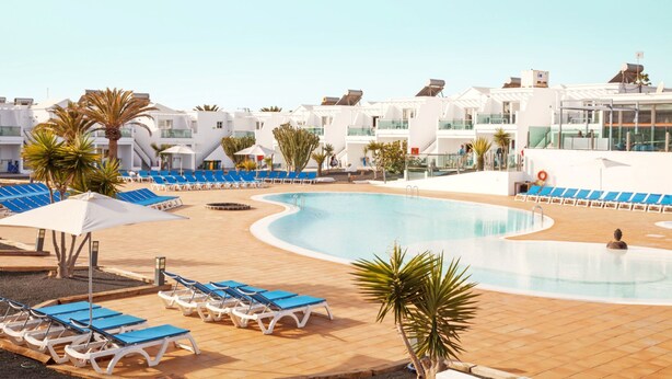 Un espace piscine avec des chaises longues bleues et des parasols, entouré de bâtiments blancs dotés de balcons.