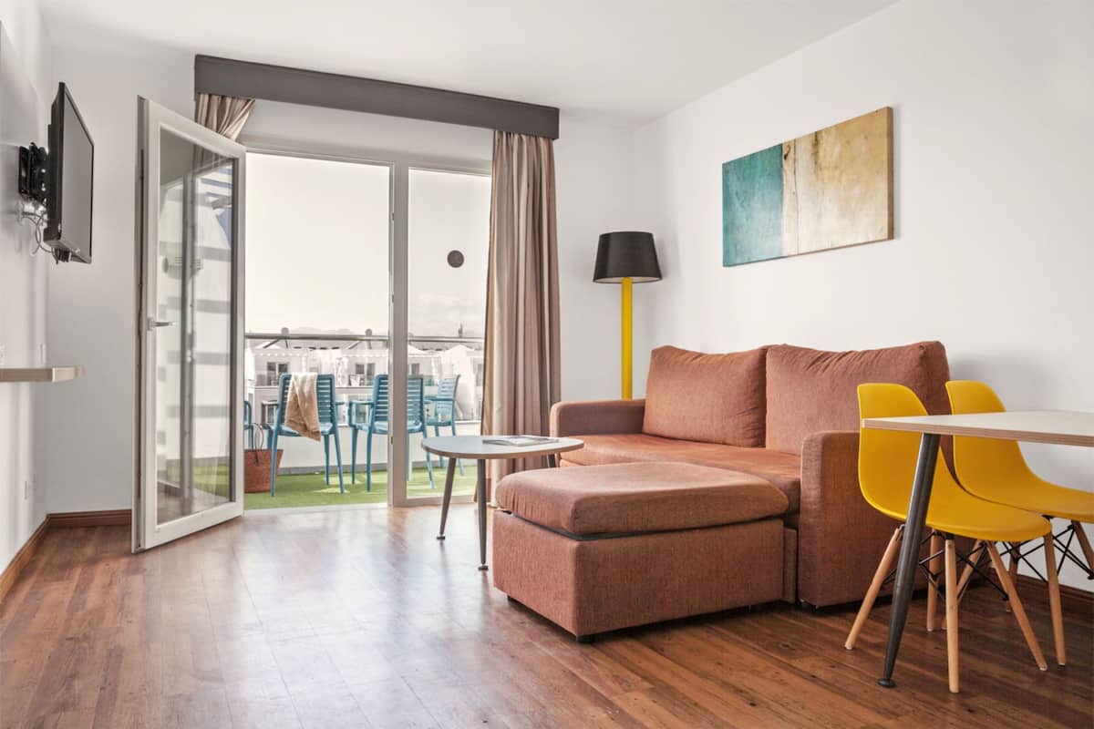 Una sala de estar moderna con un sofá, una mesita y sillas amarillas. Una puerta corrediza de vidrio da a un espacio exterior con sillas y una mesa.