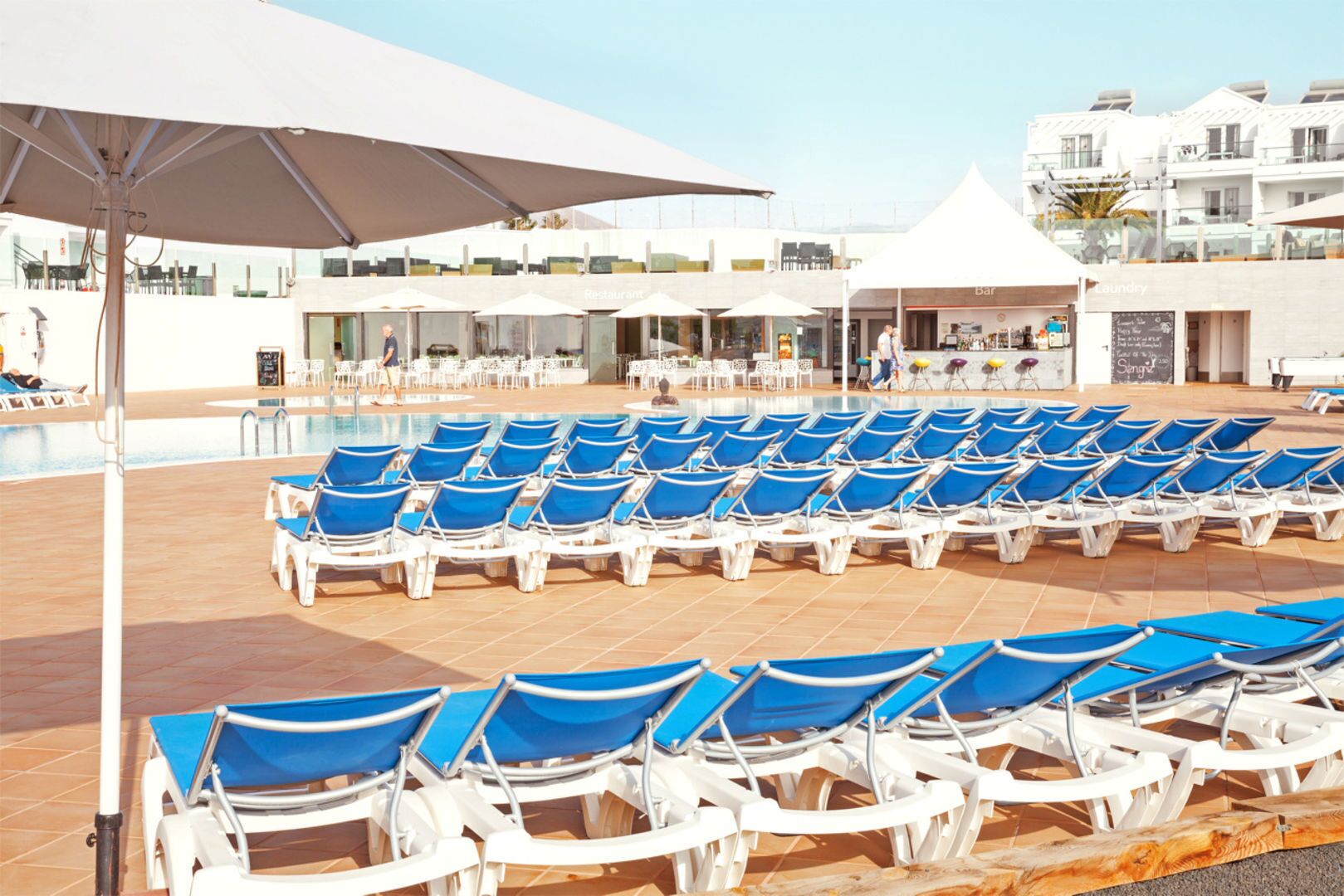 Un espace aménagé au bord de la piscine, avec des rangées de chaises longues bleues et de parasols blancs, un restaurant et un bar.
