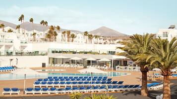A resort with a pool area, sun loungers, palm trees, and a mountain in the background.