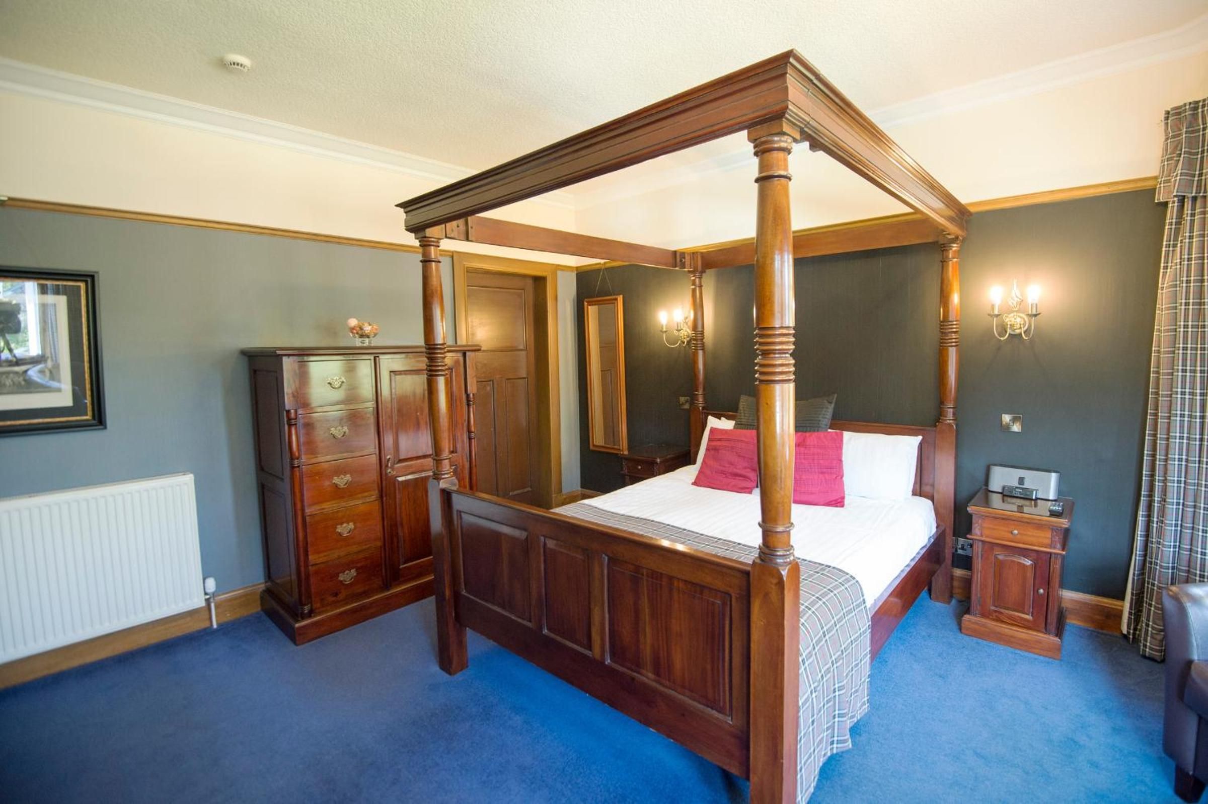 A four-poster bed with a wooden frame, a dresser with drawers, a bedside table with a lamp, and a radiator on the wall.