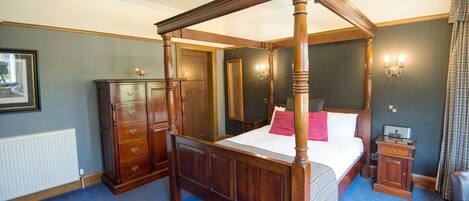 A four-poster bed with a wooden frame, a dresser with drawers, a bedside table with a lamp, and a radiator on the wall.