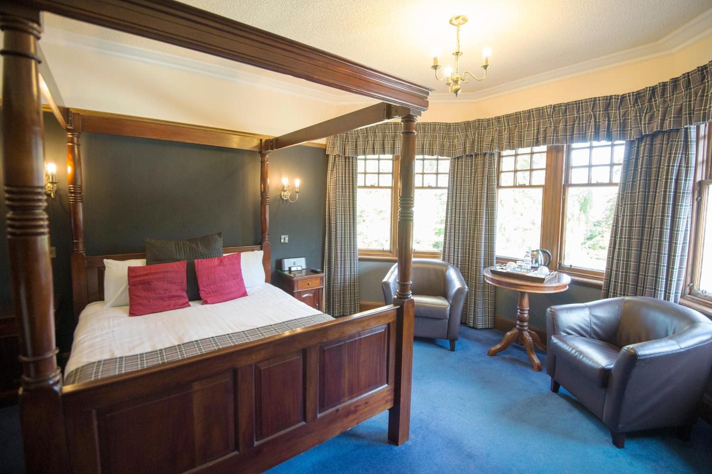 A four-poster bed with a dark wooden finish, a small round table with a tray, and a leather armchair in a room with plaid curtains and a chandelier.