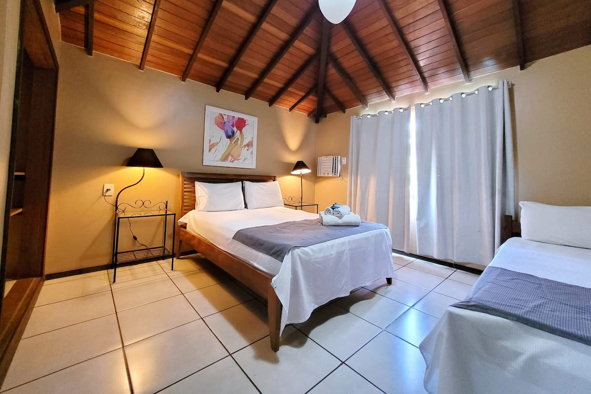 Habitación de hotel con techo de madera, una cama con ropa blanca, una mesita de noche con lámpara y una ventana con cortinas.
