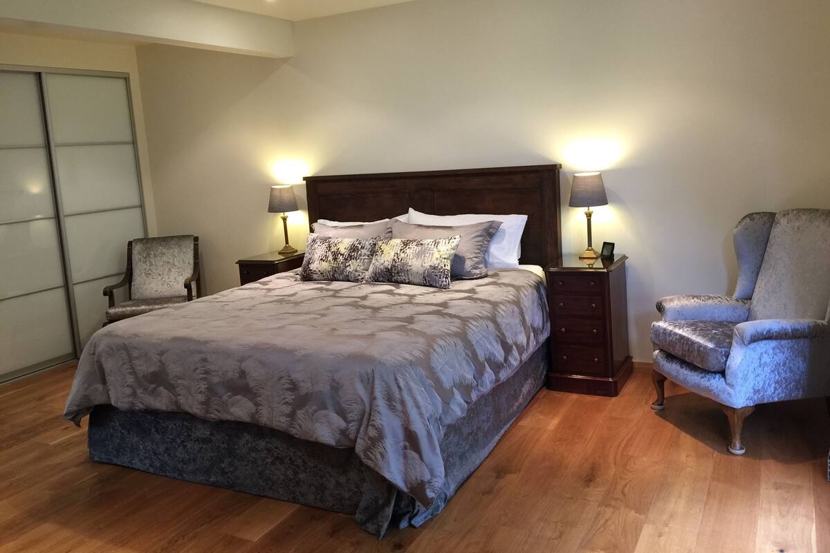 A bedroom with a large bed, two bedside tables with lamps, a chair, and a wooden floor.