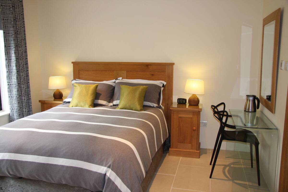 A neatly made bed with striped bedding, two bedside lamps, a glass table with a chair, and a large mirror on the wall.