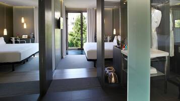 A hotel room with two beds, a desk, a bathroom, and a view of greenery outside.