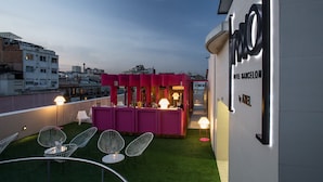 Un bar en la azotea con una zona de bar rosada, sillas blancas y vistas de la ciudad.