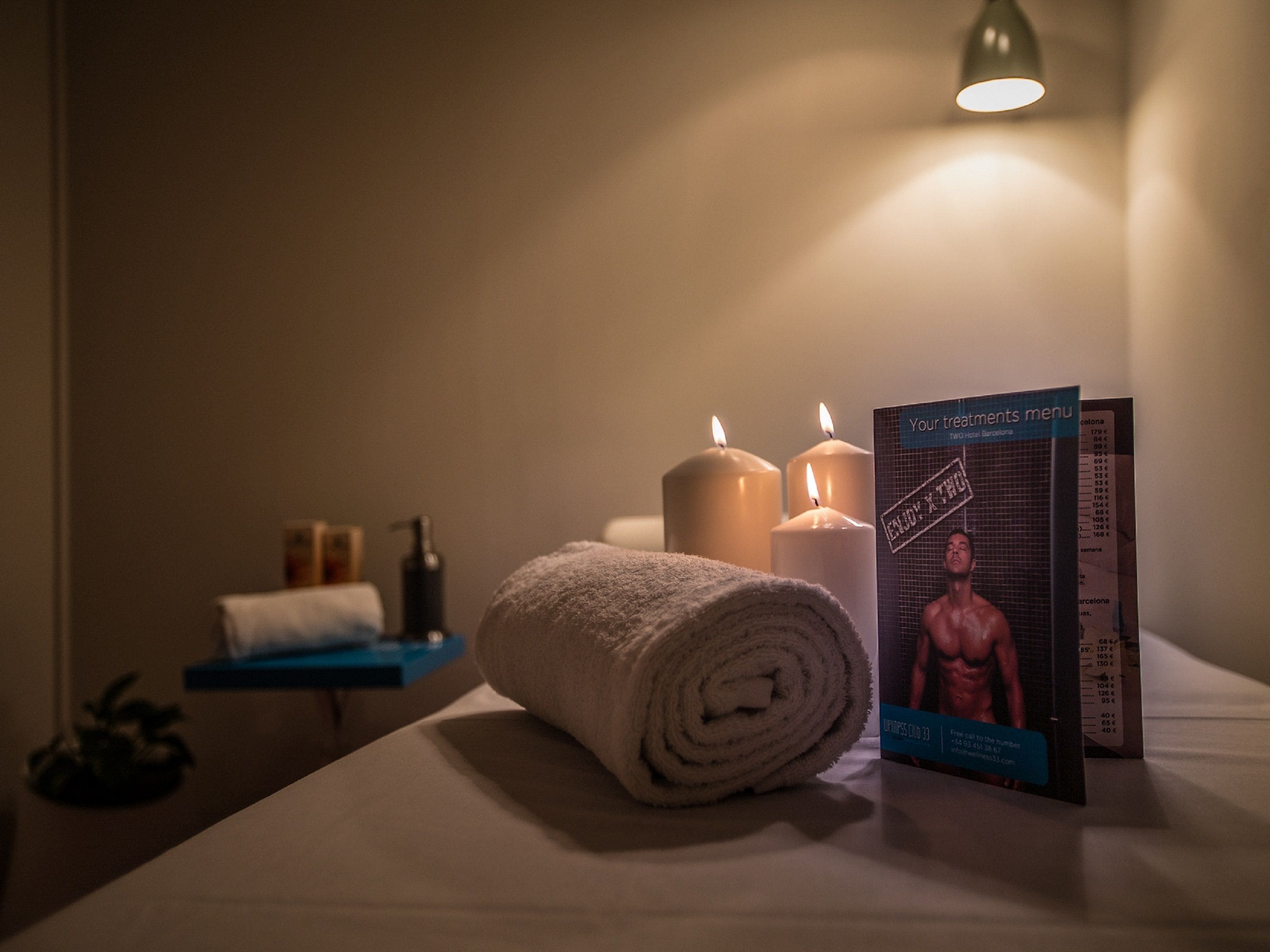 A neatly arranged room with a rolled towel, three lit candles, a menu, and a small shelf with toiletries.