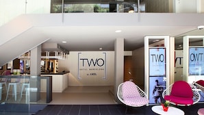 Entrada de un hotel con diseño moderno, destacando un letrero grande que dice 'TWO HOTEL BARCELONA', un área de bar y asientos con una planta en maceta.