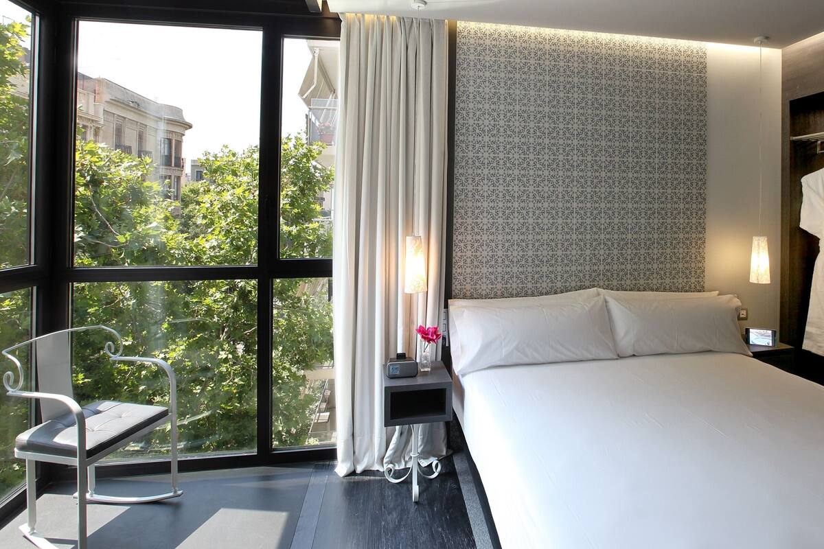 Una habitación de hotel moderna con un ventanal grande, una cama, una mesita de noche con lámpara, una bata colgada y vistas a la ciudad.