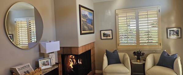 A living room with a fireplace, two armchairs, a round mirror, and a window with blinds.