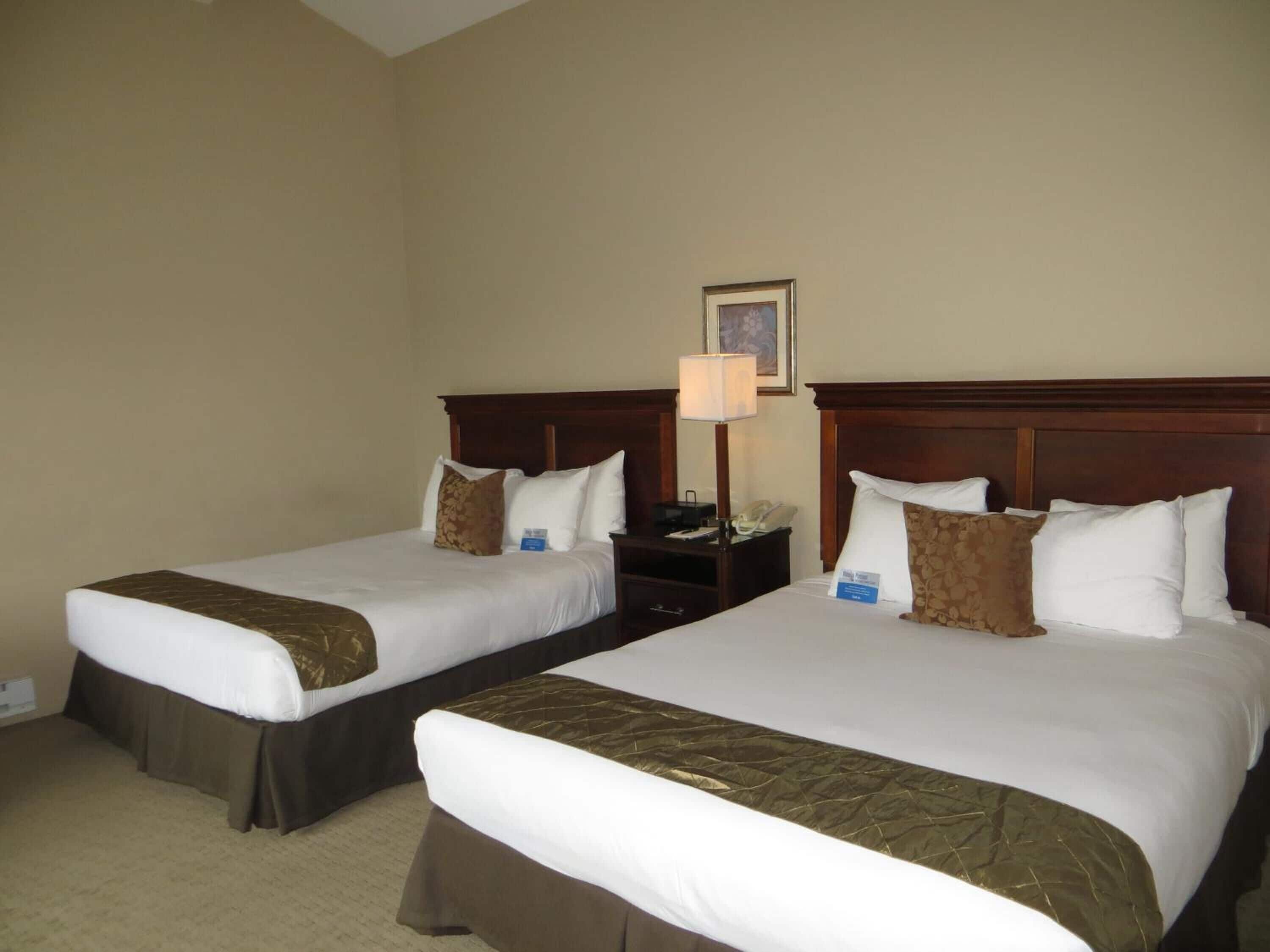 Two queen beds in a hotel room with a wooden headboard, a nightstand, a lamp, and a framed picture on the wall.