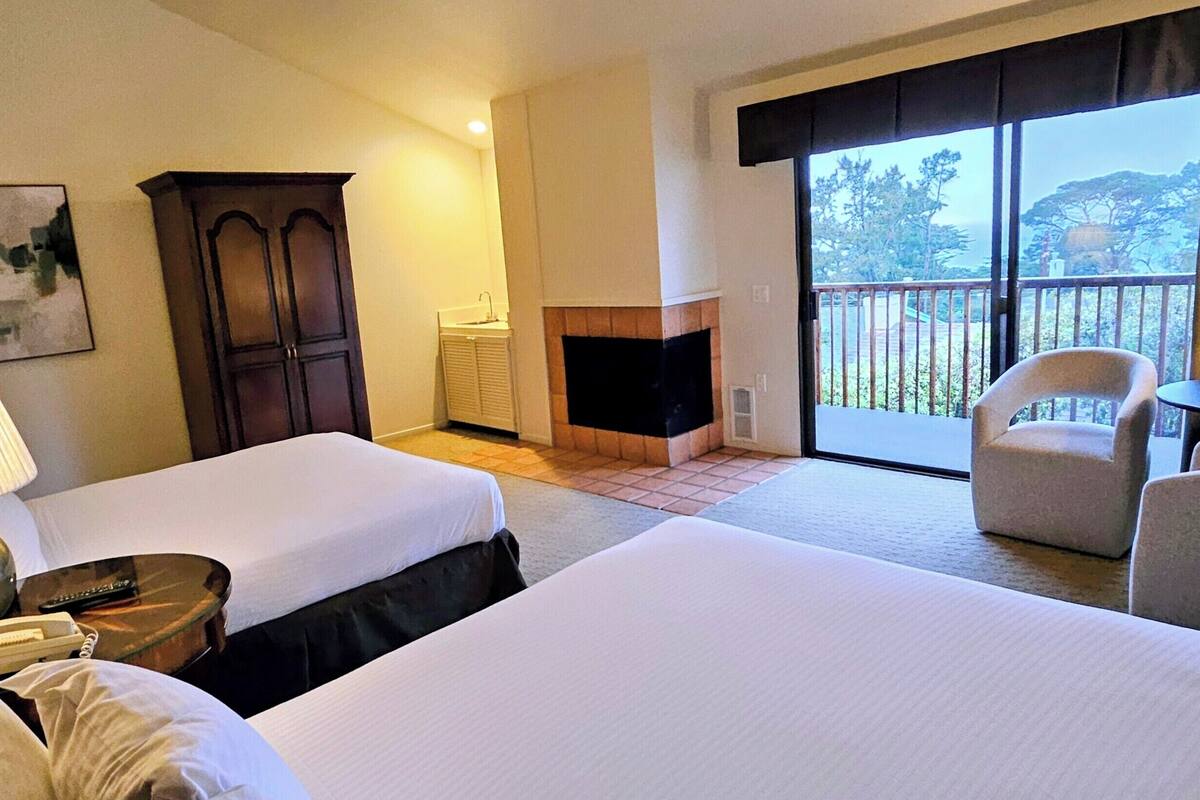 Une chambre d’hôtel avec deux lits, une cheminée, un balcon avec un fauteuil et un tableau au mur.