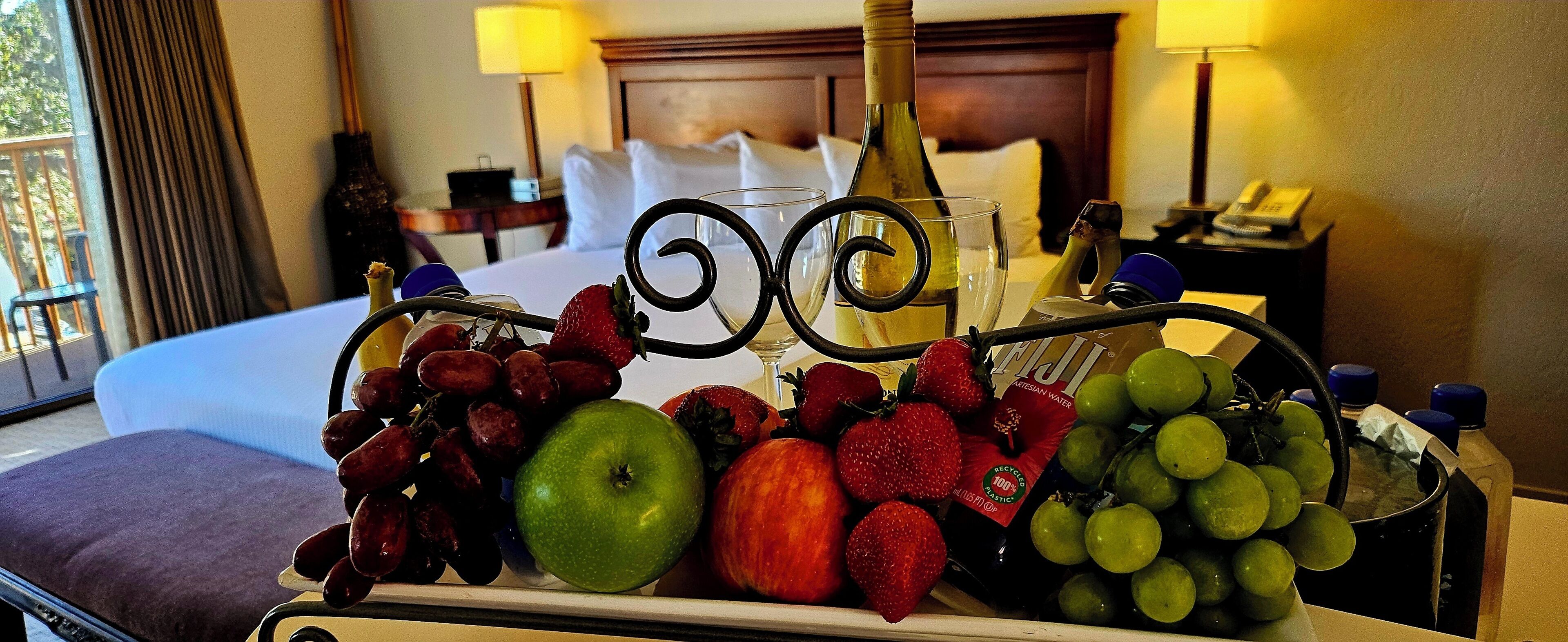 Habitación de hotel con una cama, una mesita de noche, una lámpara y una bandeja con frutas y una botella.