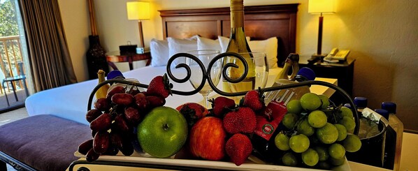 A hotel room with a bed, a nightstand, a lamp, and a tray with fruits and a bottle.
