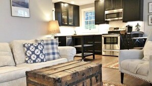 A living room with a sofa, a coffee table, and a kitchen area with dark cabinets, a microwave, and an oven. - The Parsonage Inn (Orleans)