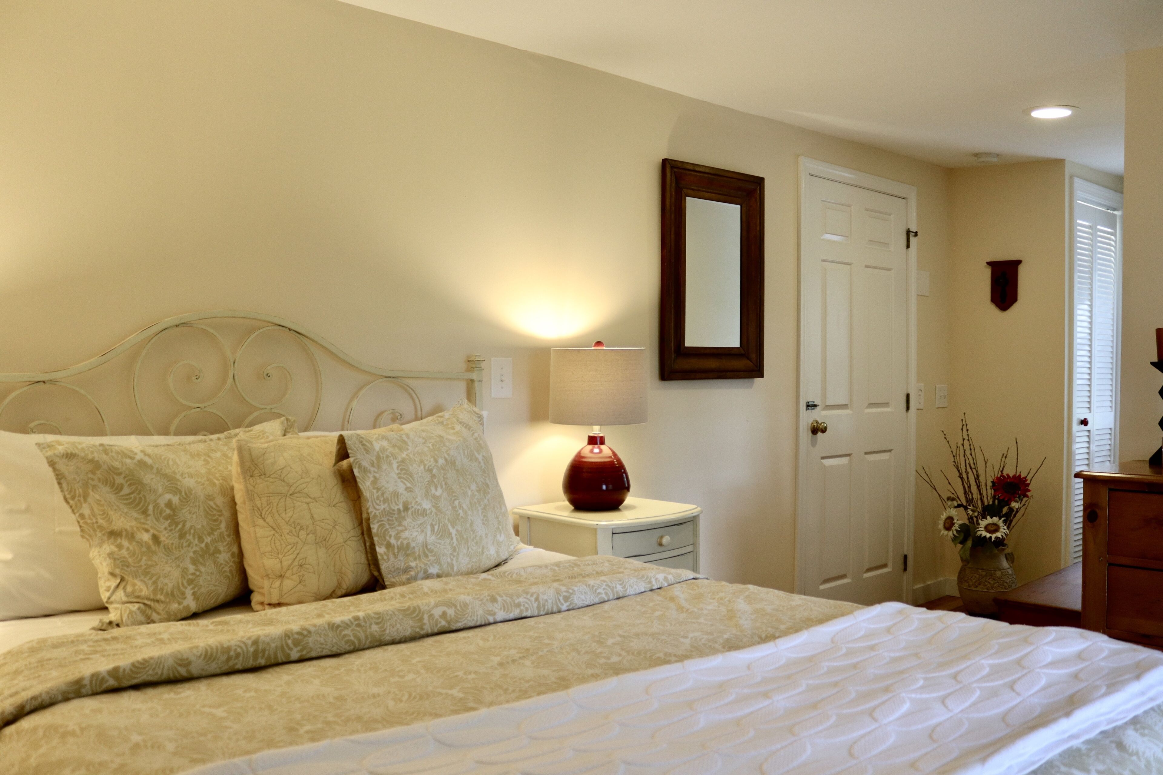 Un lit bien fait, avec une couvre-lit à motifs et des oreillers, une lampe de chevet, une table de nuit et une porte surmontée d’un miroir.