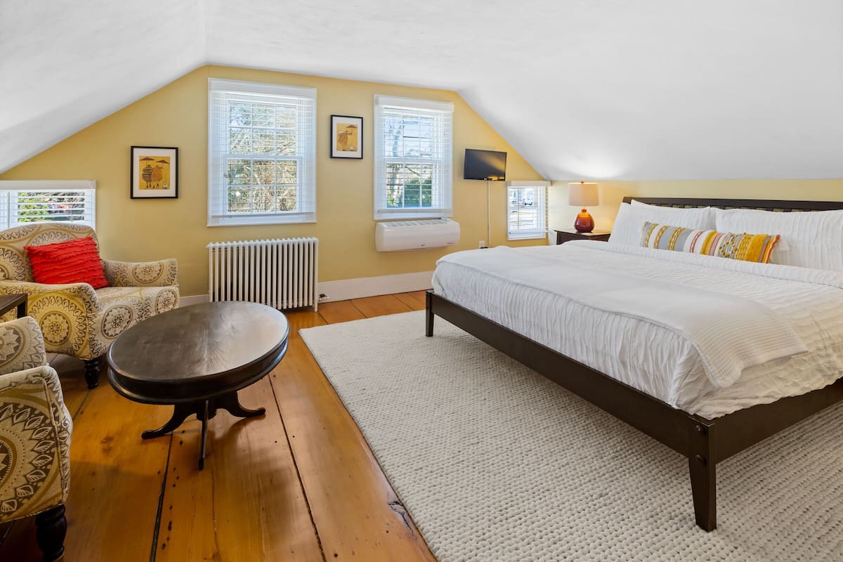 A bedroom with a large bed, a sofa, a table, and two windows with blinds.