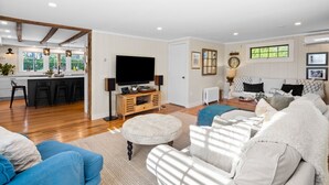 A spacious living room with a large TV, comfortable seating, a kitchen area with bar stools, and a view of the outdoors. - The Parsonage Inn (Orleans)