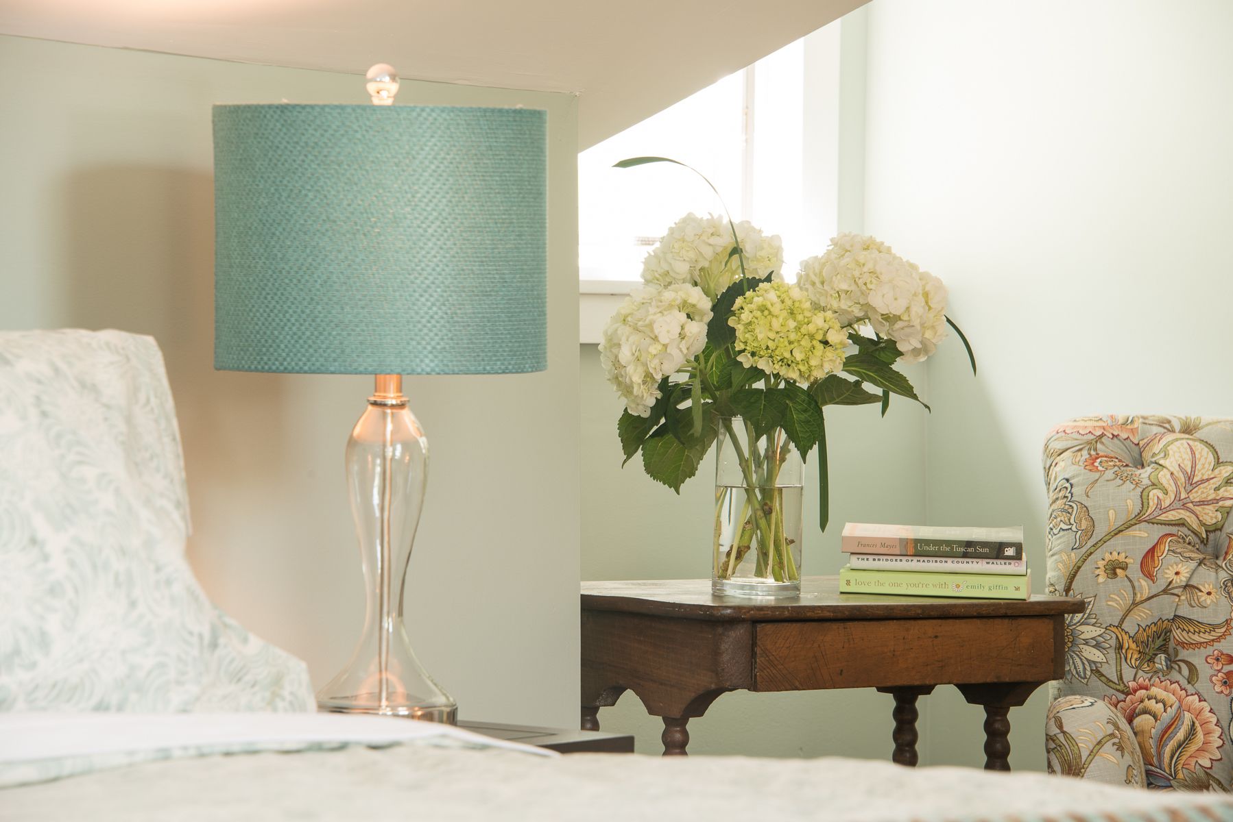 Een kamer met een bed, een lamp, een vaas met bloemen, een klein tafeltje met boeken en een gestreept fauteuil.