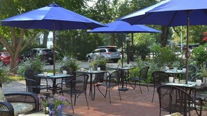 An outdoor patio with tables and chairs under blue umbrellas, surrounded by greenery and parked cars. - The Parsonage Inn (Orleans)