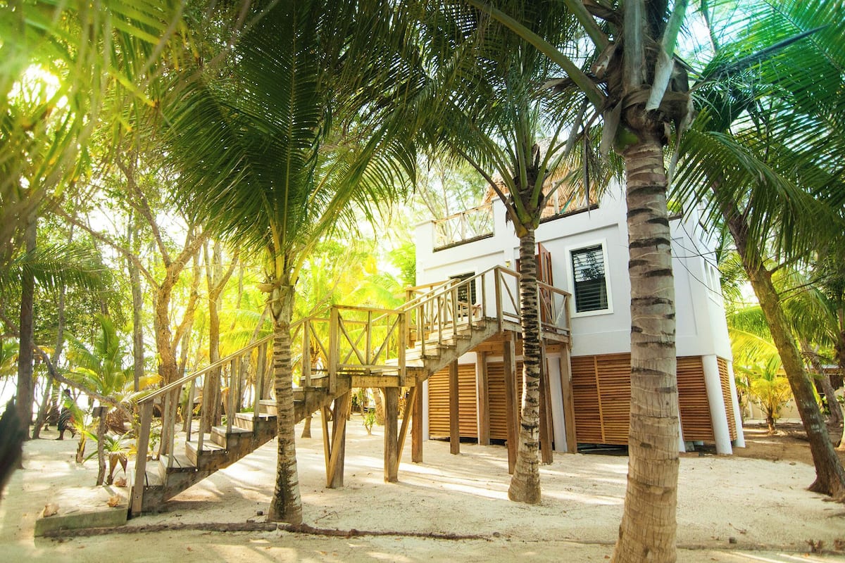 A wooden staircase leading to a white building with wooden shutters, surrounded by palm trees and sandy ground.